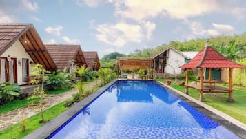 Un área de piscina con una piscina azul cristalina, rodeada de pequeñas casas con tejados rojos y una estructura con techo de paja.