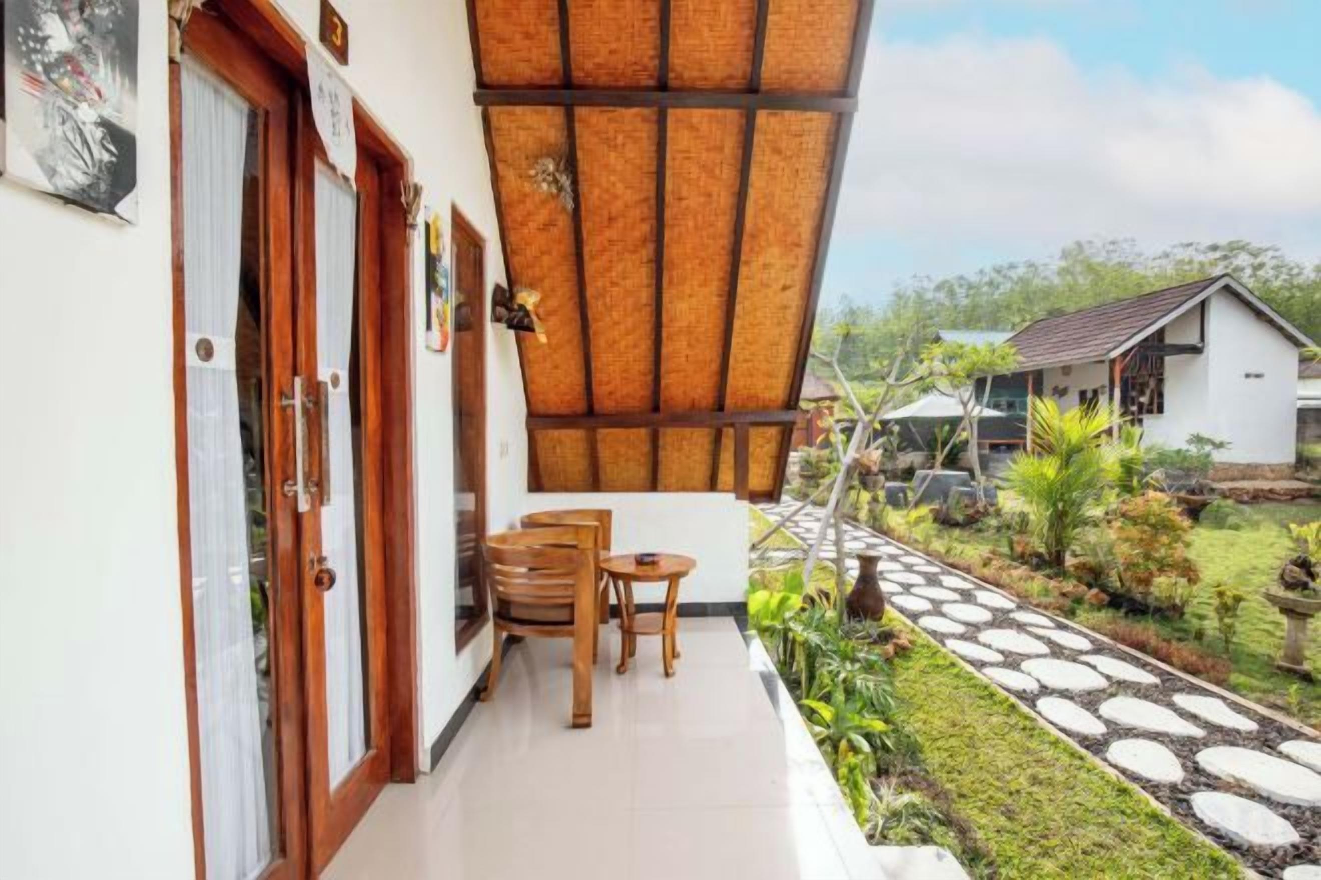 A balcony with a wooden table and chairs, a thatched roof, and a garden path leading to a white building.