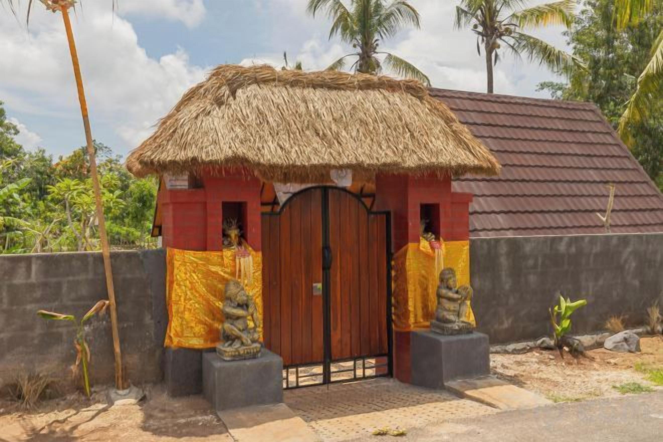 A thatched-roof structure with a gated entrance, statues, and a red wall.