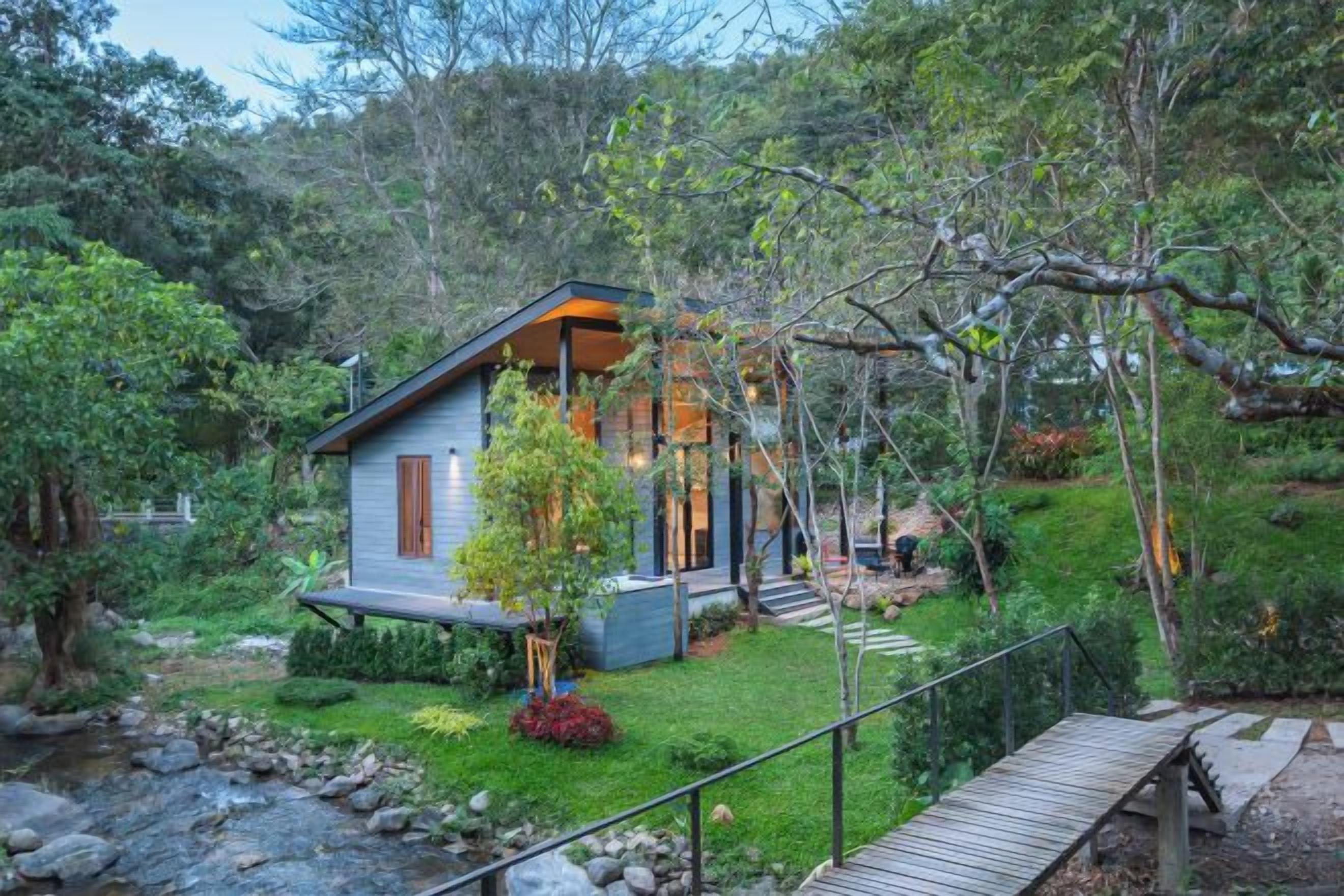 A modern house with a wooden deck, surrounded by lush greenery and a small stream.