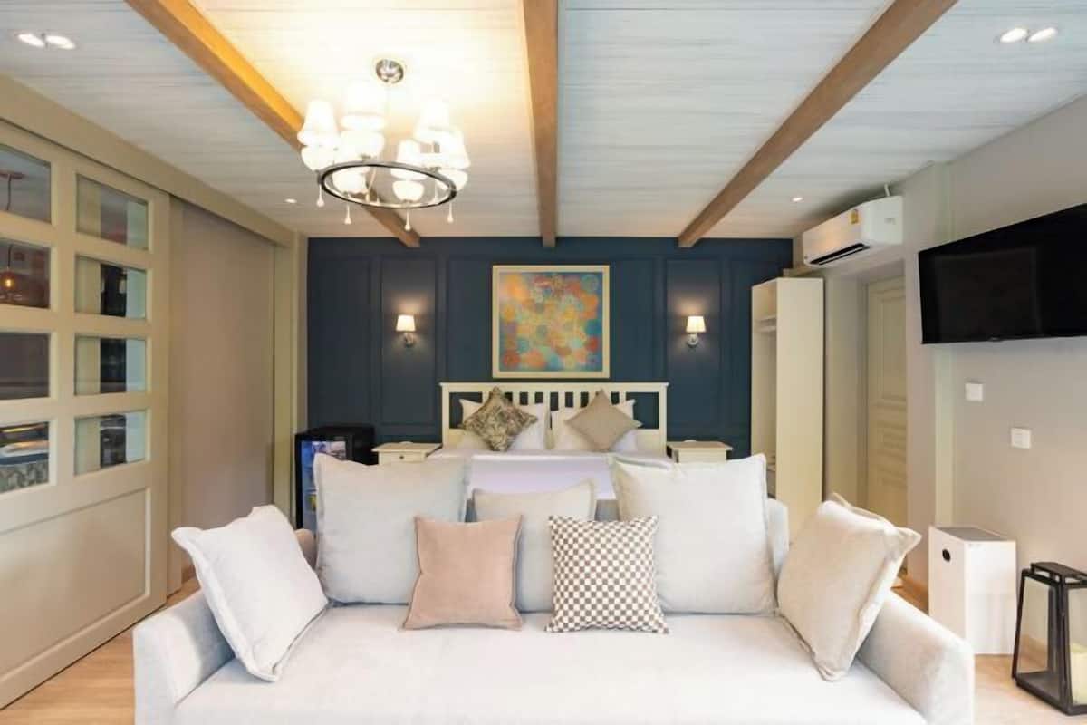 A bedroom with a bed, a white sofa, a chandelier, a painting on the wall, and a TV mounted on the wall.
