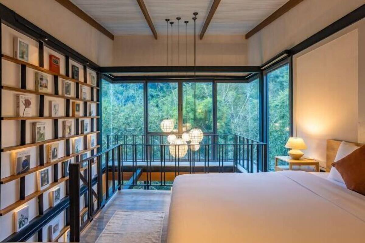 A bedroom with a large bed, a bookshelf, and a view through large windows.