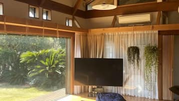 A room with a large flat-screen TV, a wooden ceiling, and a view of greenery outside.