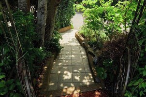A narrow stone pathway surrounded by dense greenery, leading to a paved area.