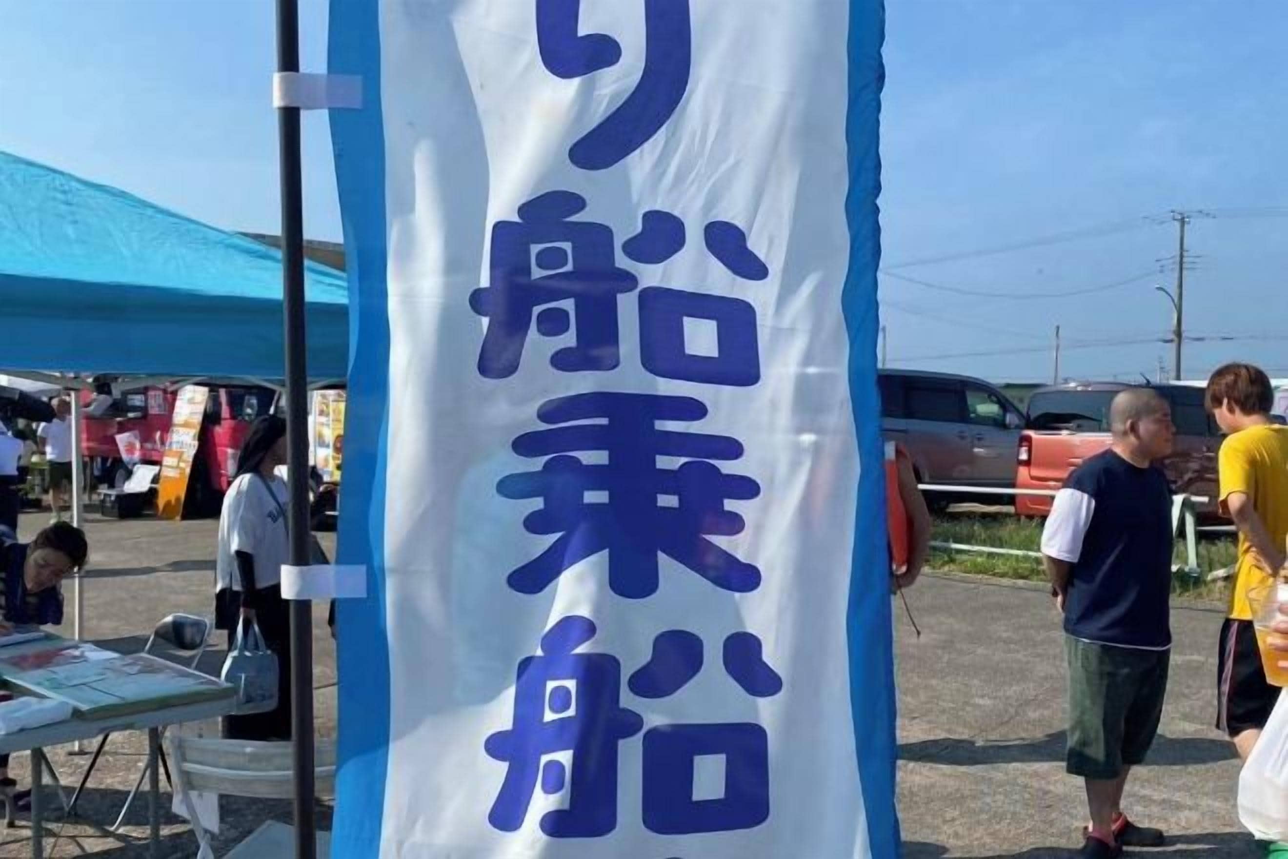 A banner with Japanese characters, a blue tent, and a parking lot with cars and people.
