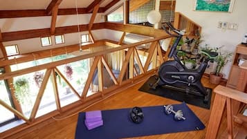 Een kamer met een houten plafond, een hometrainer, een yogamat en een raam met uitzicht op groen.