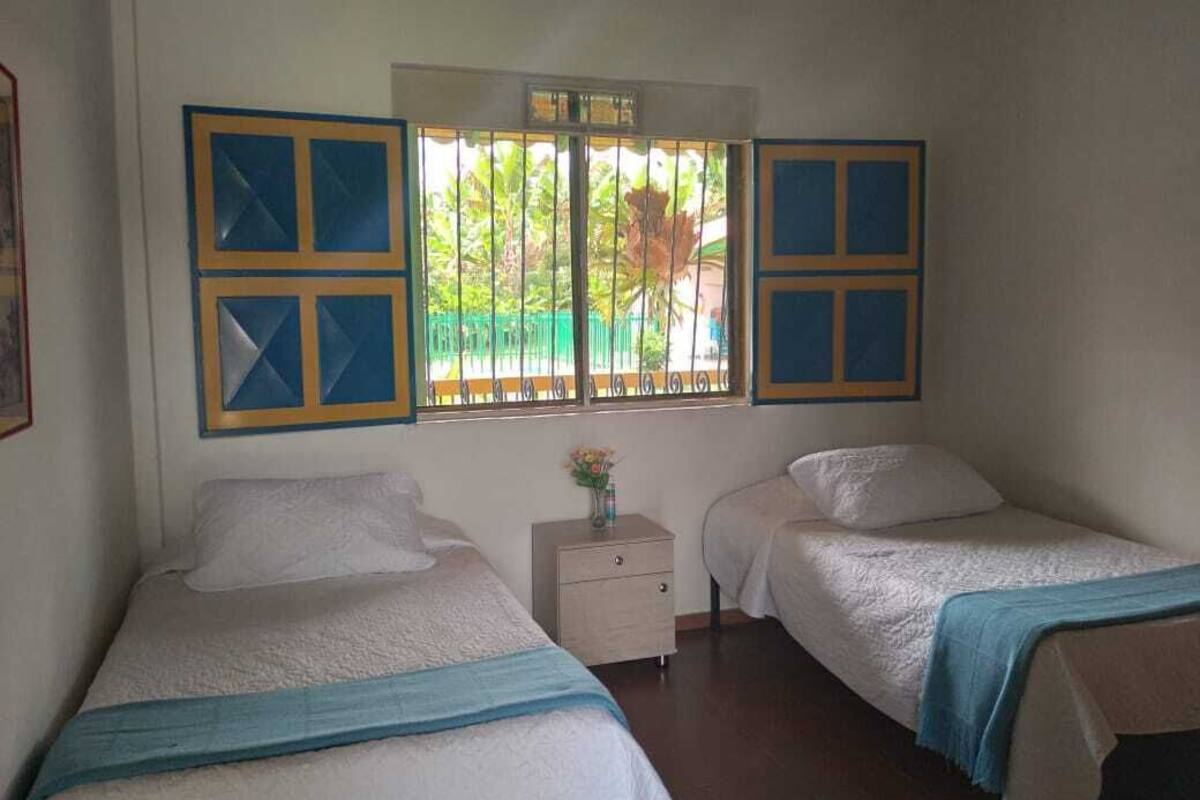 A room with two beds, a small bedside table with a vase of flowers, and a window with a view of greenery outside.