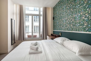 A hotel room with a bed, a nightstand, a window with curtains, and a floral-patterned wall.