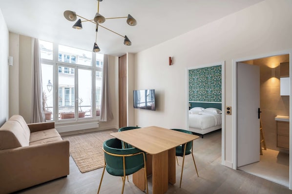 A modern hotel room with a sofa, a wooden dining table with chairs, a TV, a bed, and a view of the city.