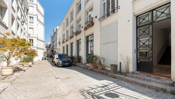 Una stretta strada acciottolata fiancheggiata da edifici a più piani, un'auto parcheggiata e vasi con piante.