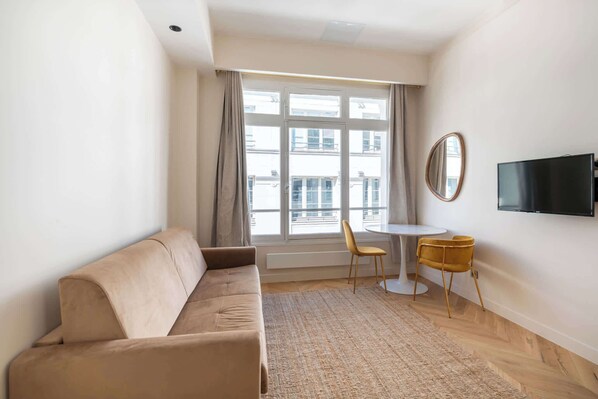 A modern living room with a sofa, a dining table with chairs, a TV mounted on the wall, and large windows with curtains.
