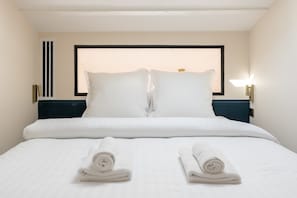 Habitación de hotel con una cama bien hecha, dos almohadas, dos toallas enrolladas, una mesita de noche con lámpara y un radiador.