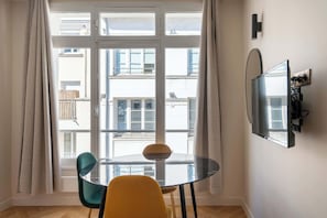 Ein modernes Zimmer mit Glastisch, zwei Stühlen, einem an der Wand montierten Fernseher und einem großen Fenster mit Vorhängen.