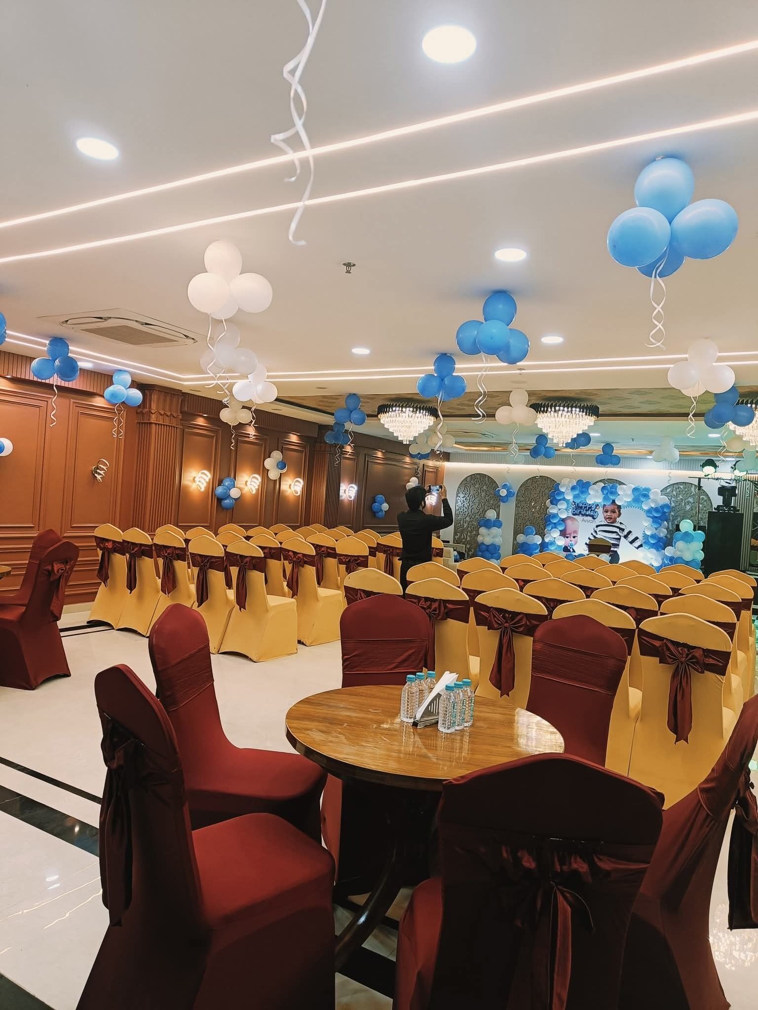 Ein Festsaal mit Stuhlreihen, einem zentralen Tisch und Deckenverzierungen, darunter Ballons und Kristalllüster.
