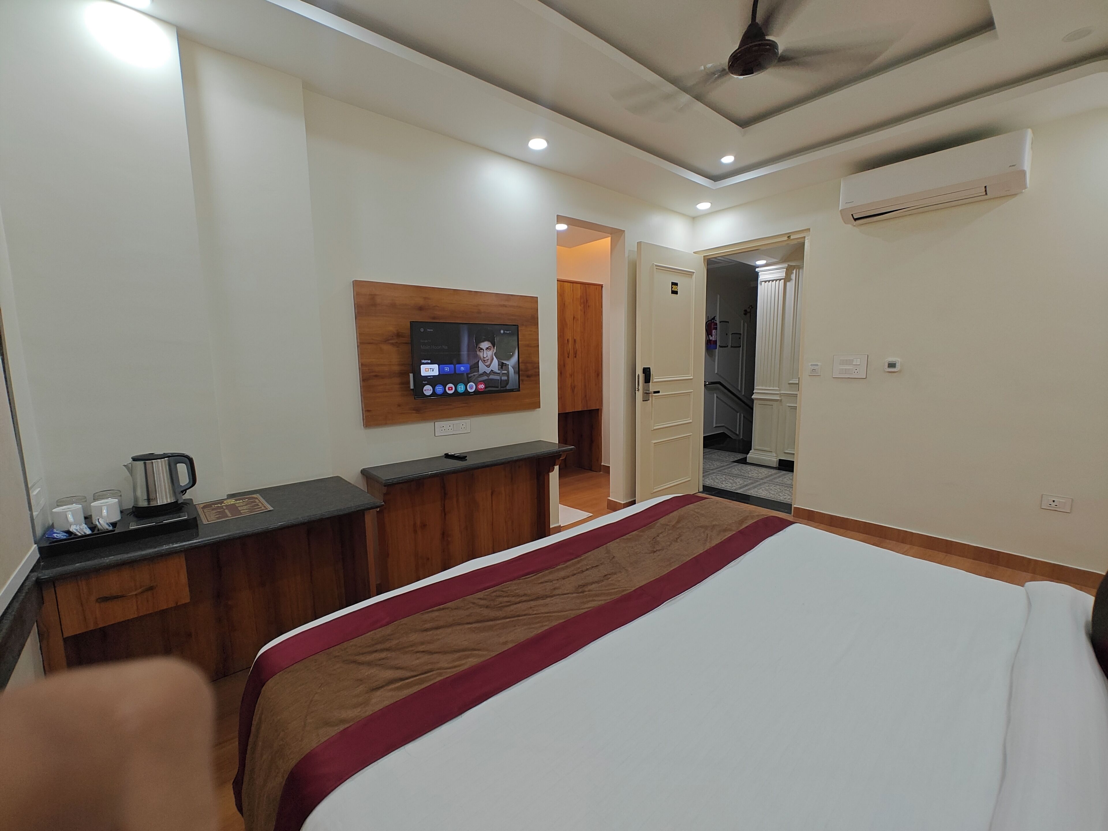 A hotel room with a bed, a desk with a coffee maker, a TV mounted on the wall, and an air conditioning unit.