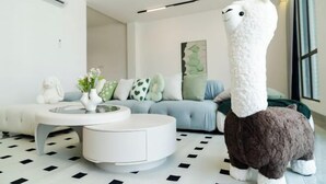 Un salon moderne avec une table basse ronde blanche, un canapé agrémenté de coussins à motifs et un grand lama en peluche.
