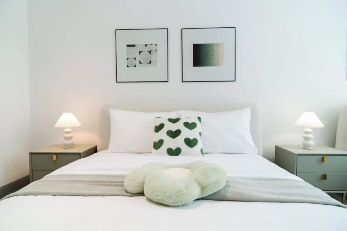 Ein ordentlich eingerichtetes Hotelzimmer mit einem Bett, zwei Nachttischen mit Lampen, einem dekorativen Kopfkissen mit Herzmustern und zwei gerahmten Kunstwerken an der Wand.
