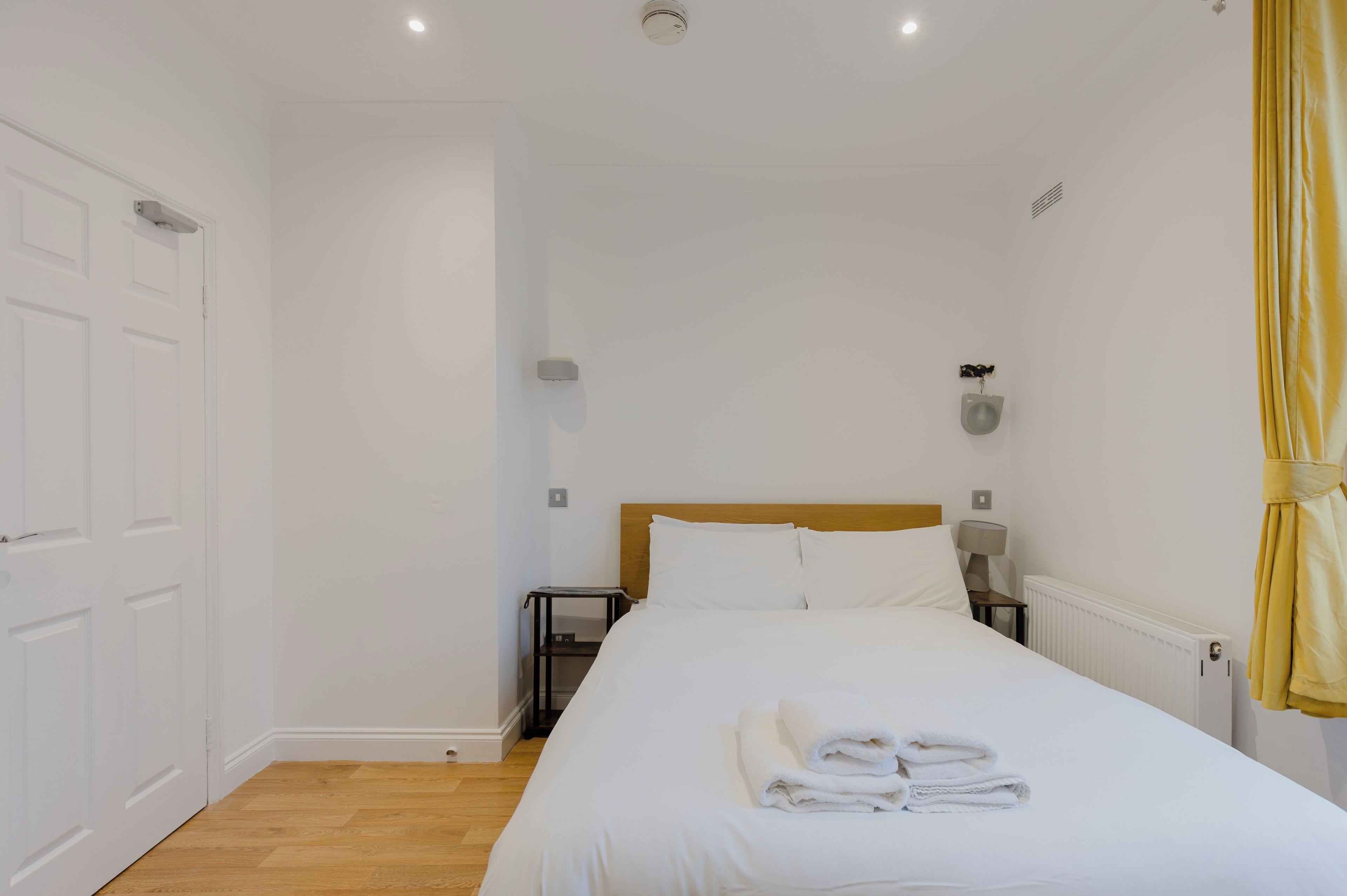 A hotel room with a bed, a bedside table, a lamp, a radiator, and a curtain.