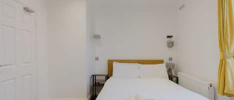 Quarto de hotel com cama, mesa de cabeceira, abajur, radiador e cortina.