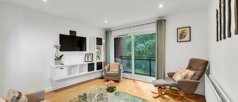 A modern living room with a glass coffee table, a sofa, a chair, a TV mounted on the wall, and a large window with a view of greenery.