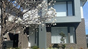 Una casa moderna de dos pisos amb façana de maó, grans finestres i un jardí ben cuidat amb arbres en flor.