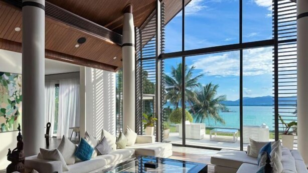 A modern living room with a large glass window offering a view of the ocean and palm trees. - 6BR Oceanfront Luxury Villa 30m Infinity Pool Villa Serenity Warm Plus (Phuket)