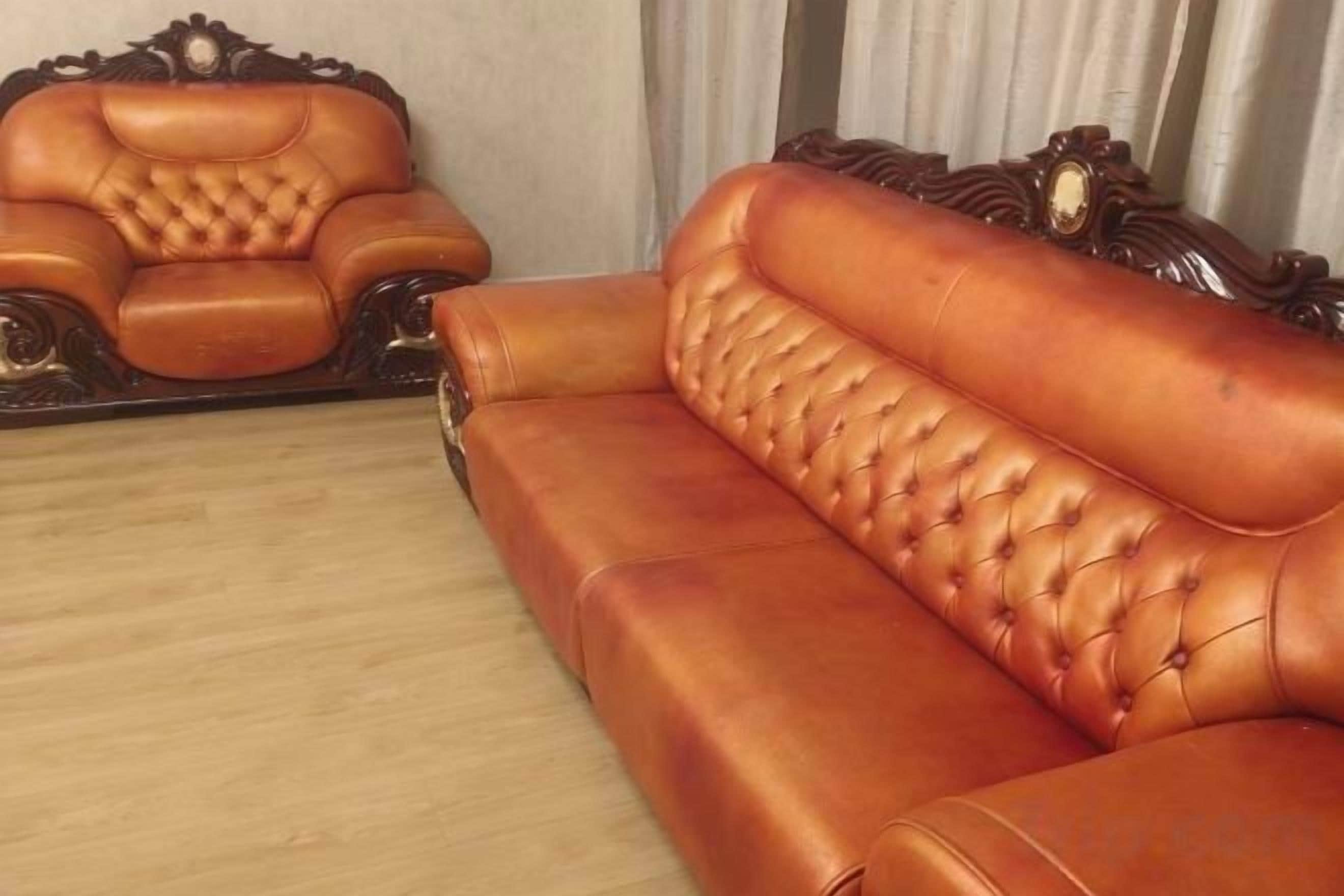 A brown leather sofa with a tufted backrest and armrests, positioned in a room with a wooden floor and a curtain in the background.