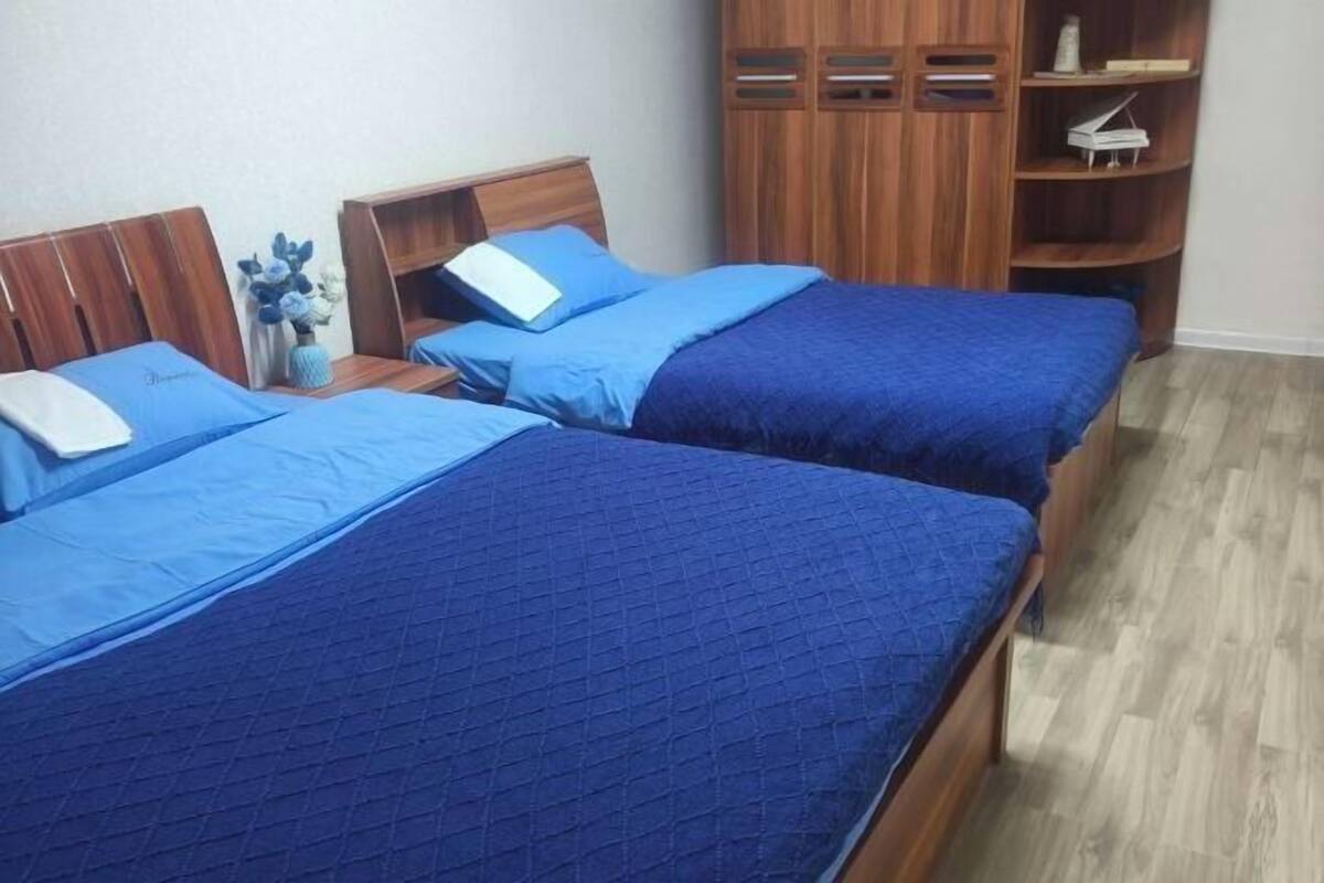 A room with two beds, each with a blue quilted bedspread and matching pillows. There is a wooden wardrobe with shelves and a piano on the right.