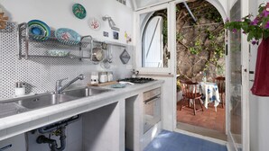 A kitchen with a sink, stove, and cabinets, a dining area with a table and chairs, and a view of a rocky garden through an open door. - Respiro del Mare (Sorrento)