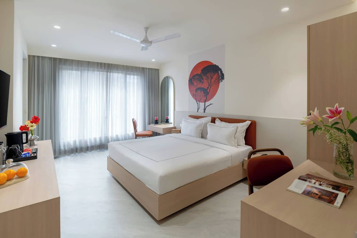 Ein Hotelzimmer mit einem großen Bett, einem Schreibtisch mit Computer, einem Stuhl, einem Fernseher und einem Fenster mit Vorhängen.
