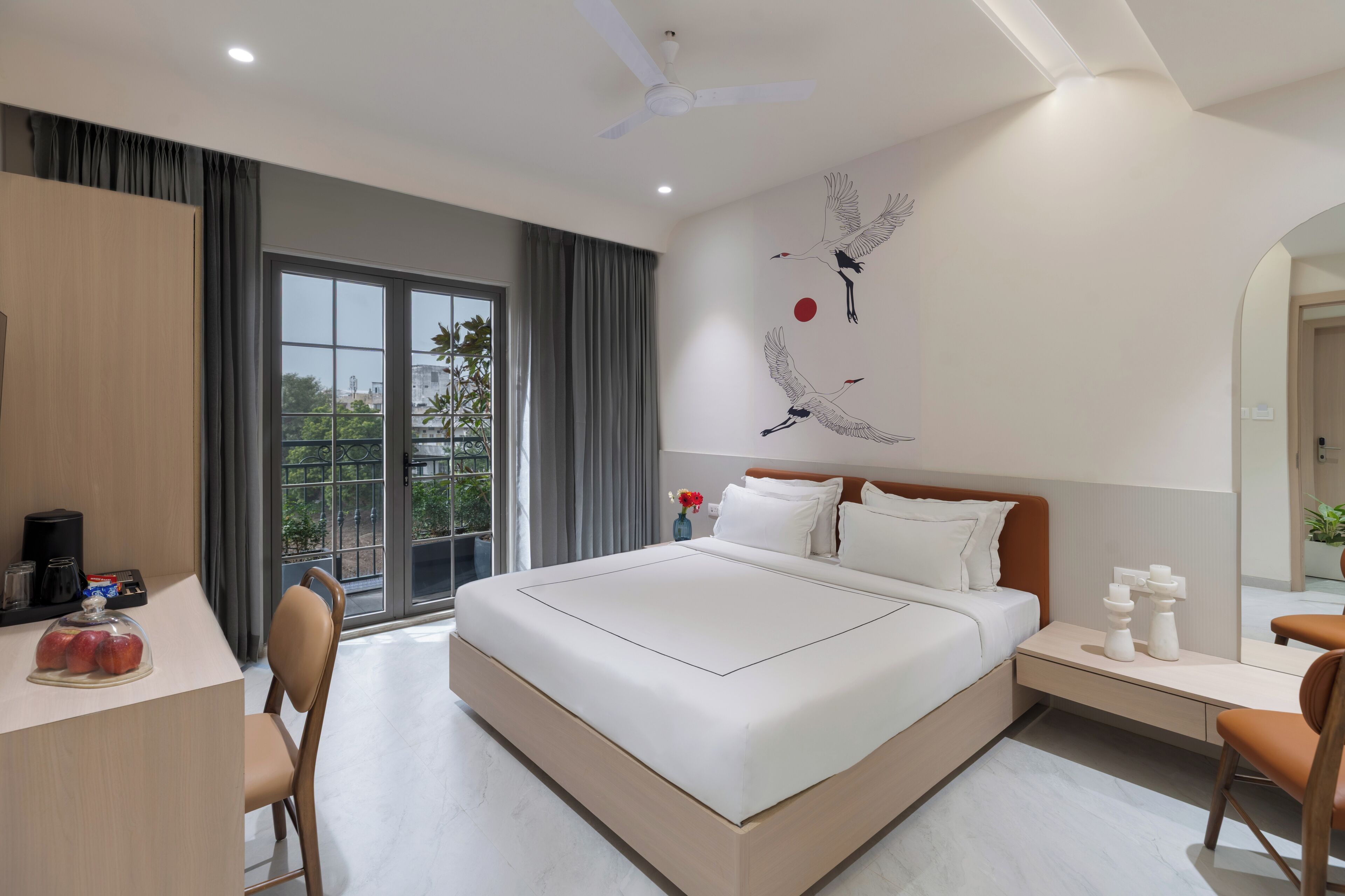 Una habitación de hotel moderna con una cama grande, un escritorio con ordenador, una silla, una mesita de noche y un amplio ventanal con cortinas.