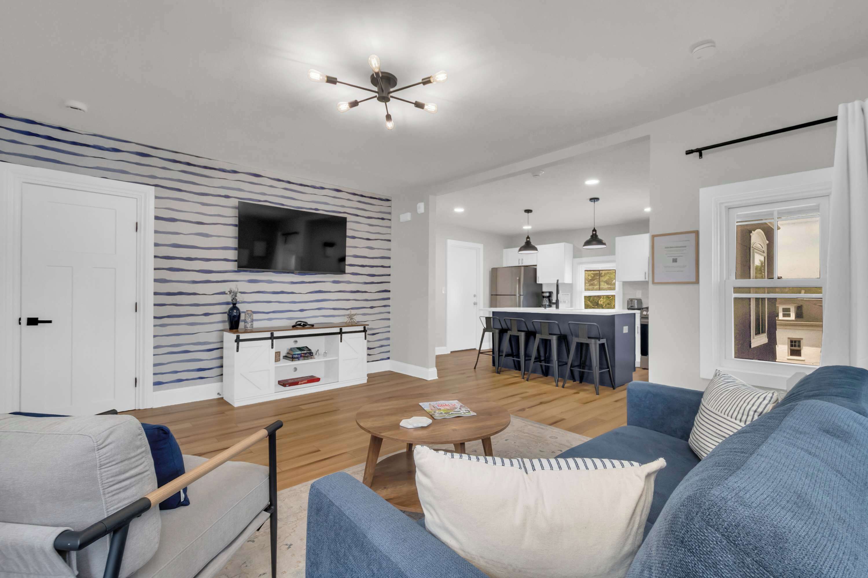A modern living room with a sofa, a coffee table, and a TV mounted on the wall. There is a kitchen area in the background with a bar and stools.