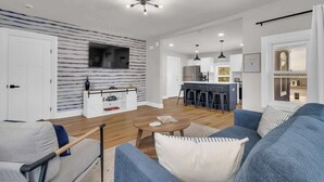Un salon moderne avec un canapé, une table basse et un téléviseur fixé au mur. On aperçoit à l’arrière-plan un coin cuisine avec un bar et des tabourets.