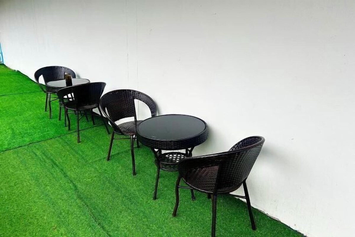 A row of black wicker chairs and a round table against a white wall, with green artificial grass in front.