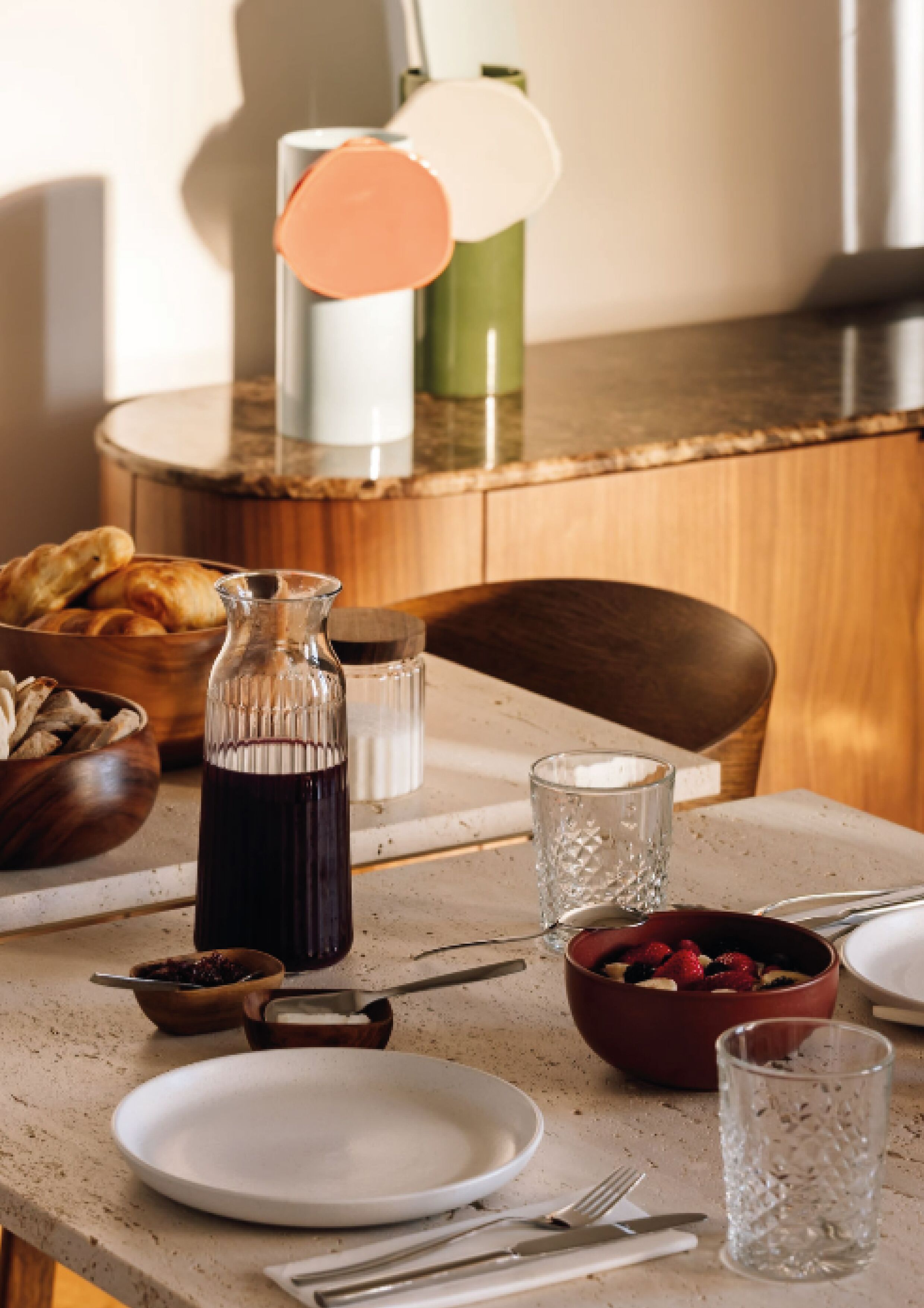 A dining table set with plates, cutlery, a glass, a bowl of fruit, a bottle, and a basket of bread.