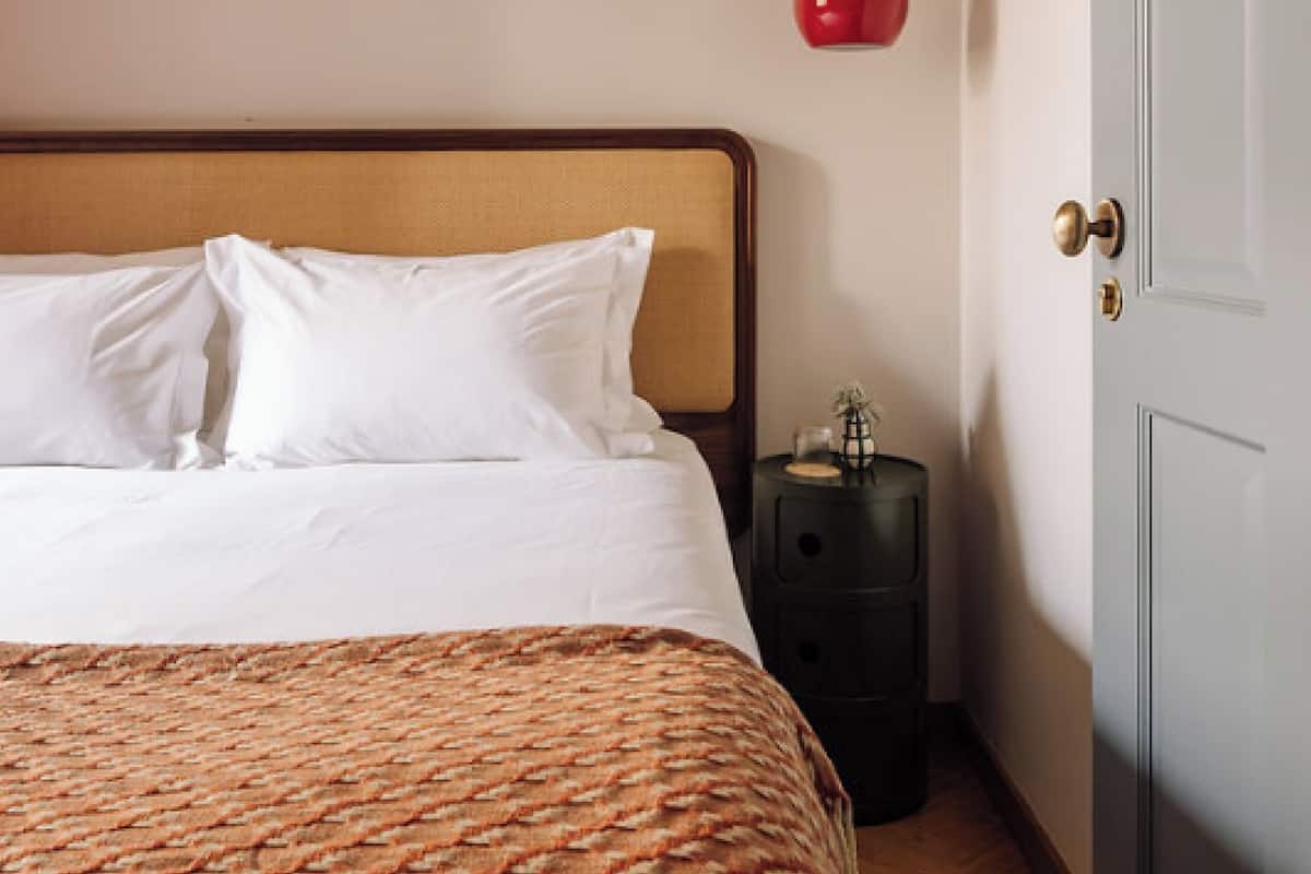 Ein ordentlich gemachtes Bett mit weißen Leinen und einer gemusterten Decke, ein kleiner Nachttisch mit einer Lampe und eine Tür rechts.