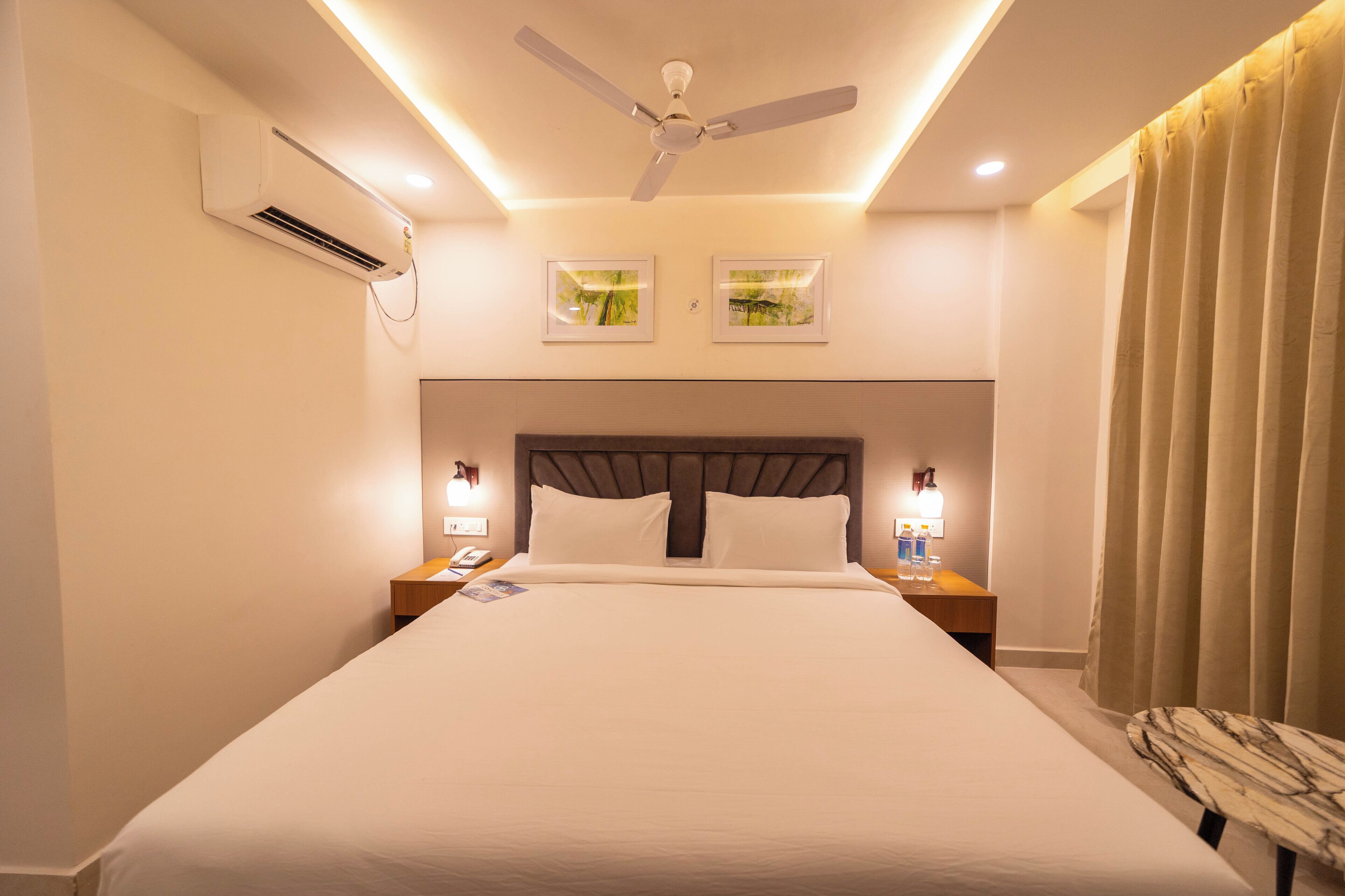 A hotel room with a large bed, a ceiling fan, wall-mounted air conditioning, two framed pictures, bedside lamps, and a curtain.