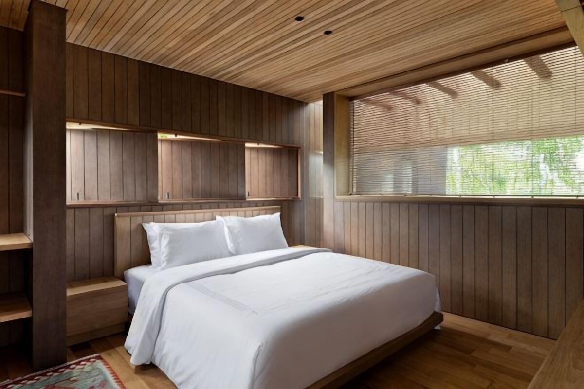 Une chambre avec un plafond en bois, des étagères intégrées, un lit avec du linge de lit blanc et une fenêtre avec des stores.