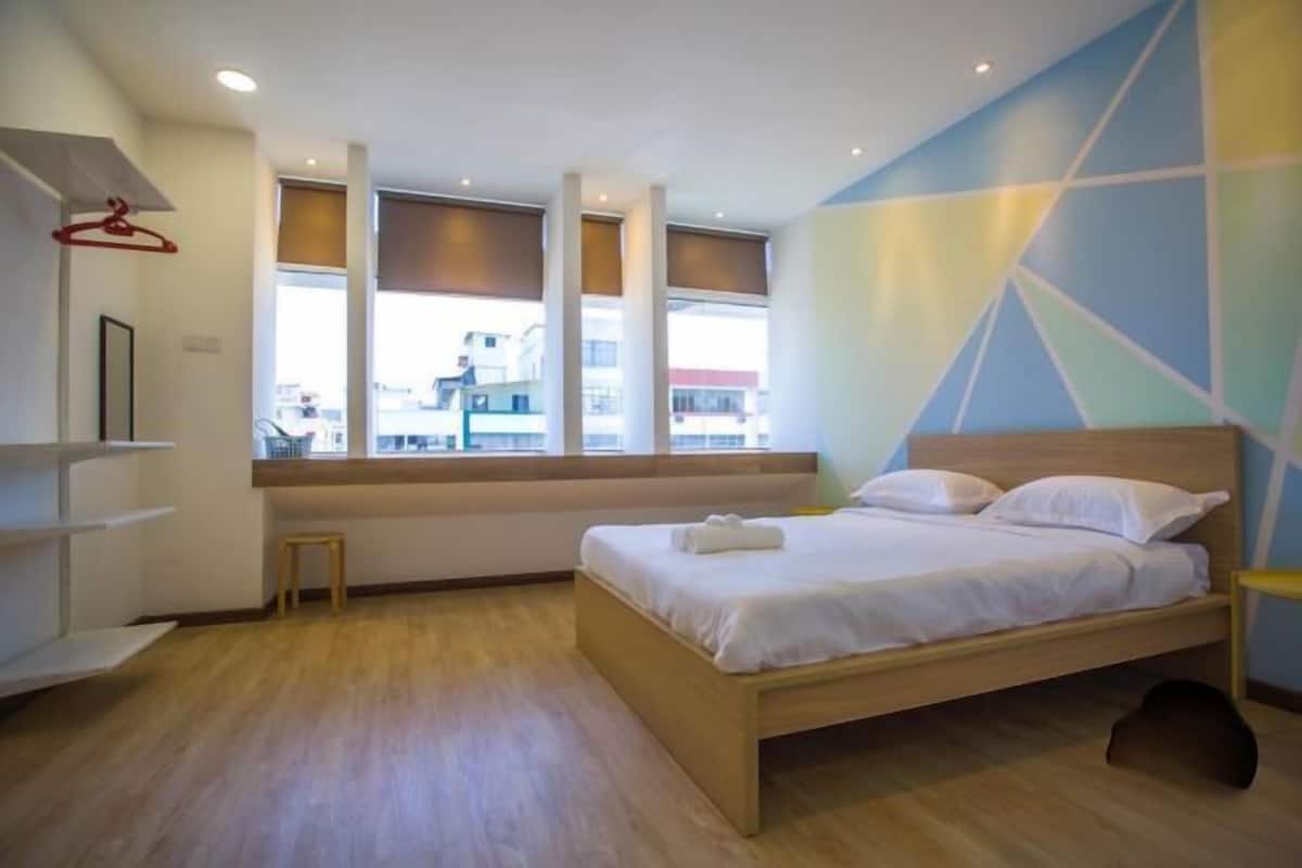 A modern bedroom with a wooden bed, a window with blinds, a small stool, and a wall with geometric patterns.