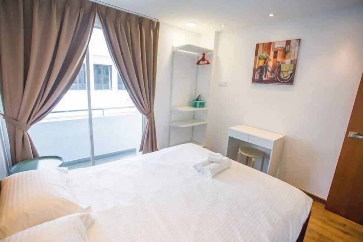 A hotel room with a bed, a window with curtains, a wall-mounted shelf with a red hanger, and a painting.