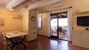 Un coin repas avec une table et des chaises en bois, un téléviseur posé sur un meuble, et une porte-fenêtre coulissante donnant sur une terrasse extérieure.