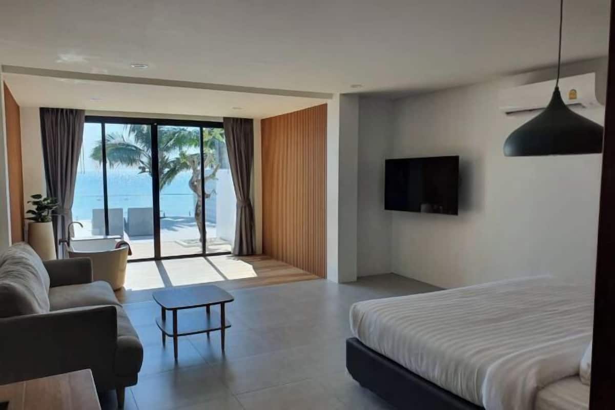 A modern hotel room with a bed, a TV, a sofa, a small table, and a balcony with a view of the sea.