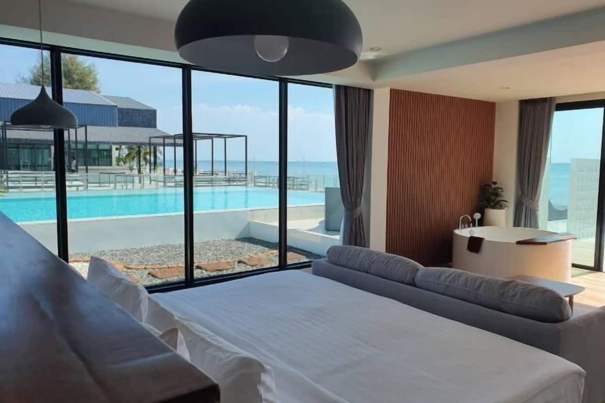 A modern hotel room with a large bed, a seating area, a small table, and a view of an infinity pool.
