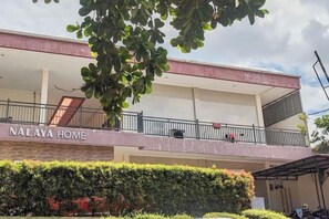 Un edificio de dos pisos con balcones, un letrero que dice 'NALAYA HOME' y un seto cuidado al frente.