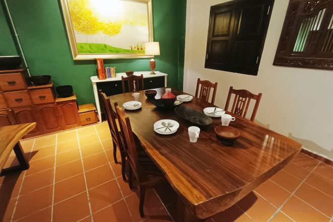 A dining area with a wooden table set for four, surrounded by chairs, with a green wall and a window in the background.