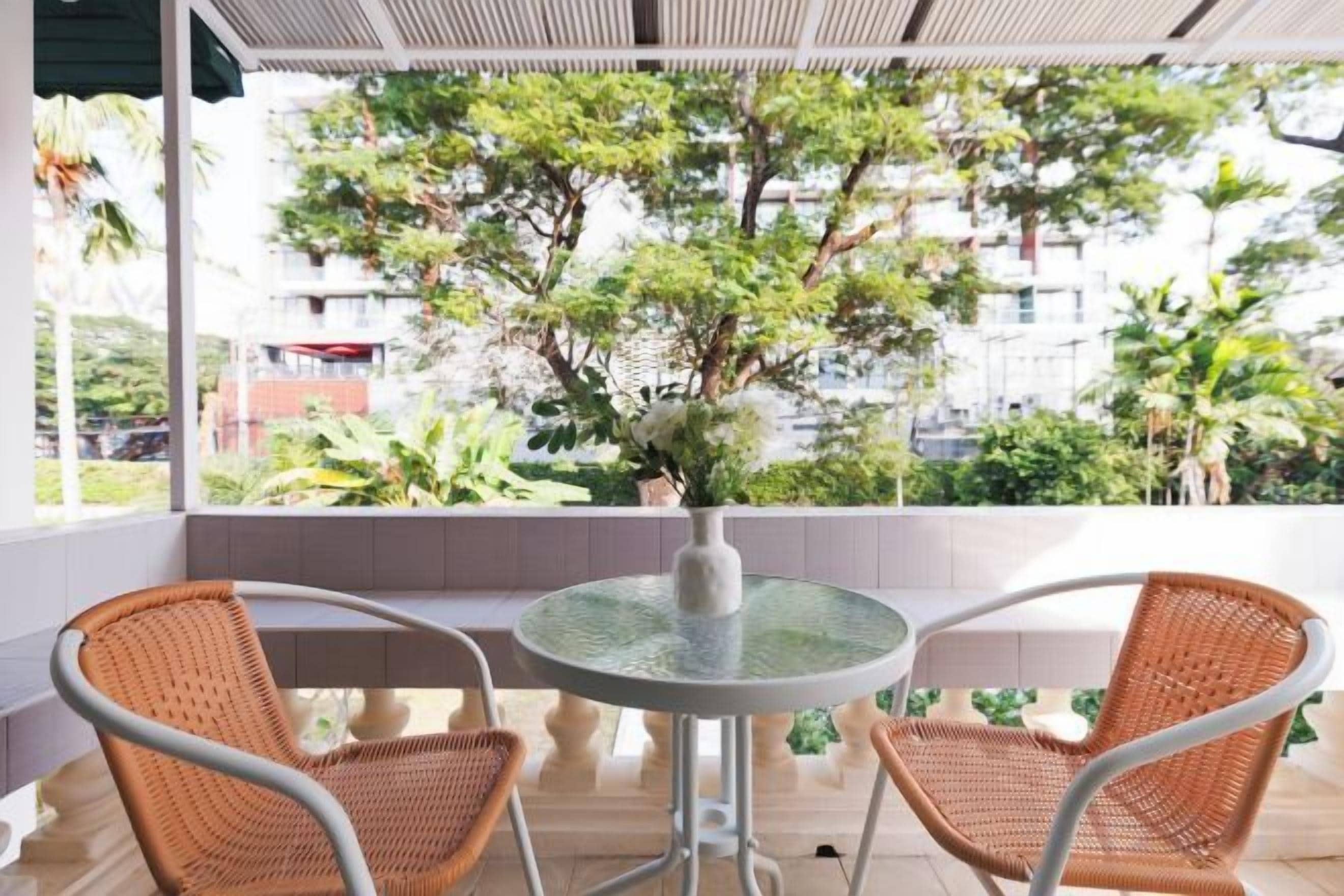A balcony with a round table and two wicker chairs, overlooking a garden with trees and a building in the background.