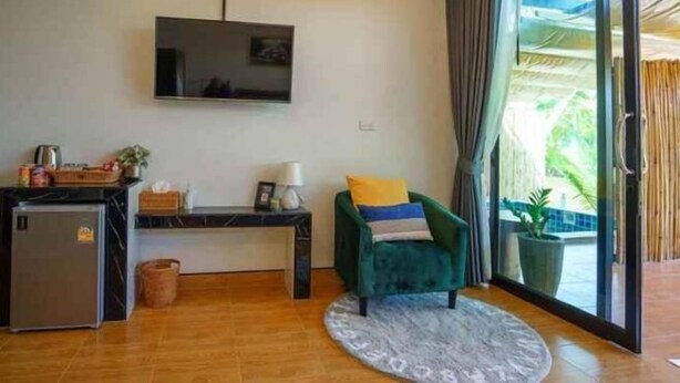 A modern living room with a flat-screen TV, a small fridge, a desk with a lamp, a green armchair, and a large window with a view of greenery. - Phetra Pool Villas (Surat Thani)