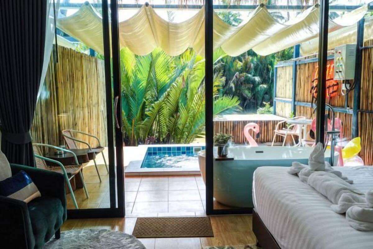 A room with a bed, a small table, a chair, a view of a pool, and a tropical garden outside.