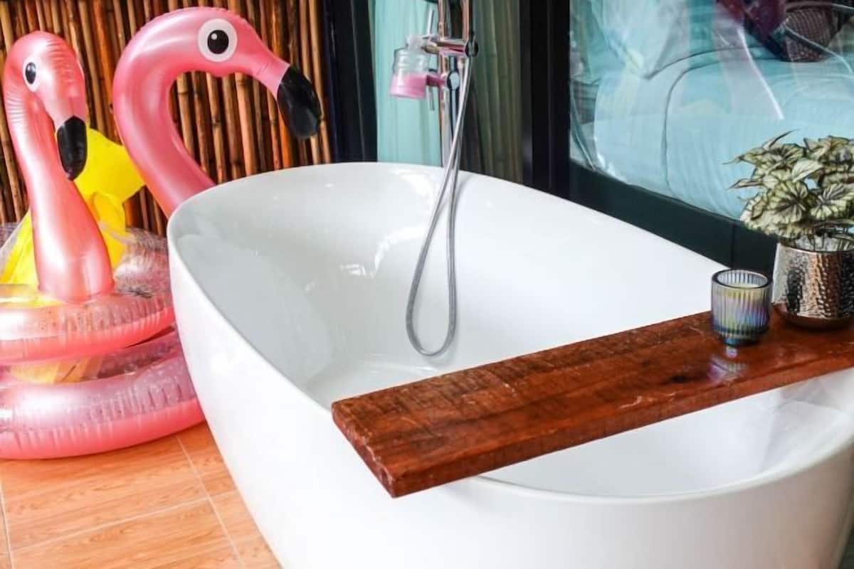 A white bathtub with a wooden tray, a pink flamingo inflatable, and a potted plant.
