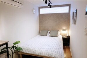 Ein Schlafzimmer mit Bett, Nachttisch, Lampe, Wanduhr und einem Fenster mit Jalousien.